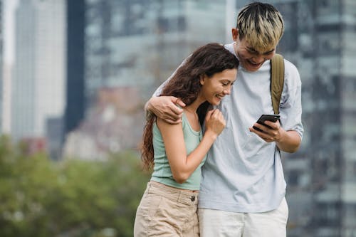 Free Positive multiracial couple embracing and laughing while sharing cellphone near urban buildings in summer Stock Photo