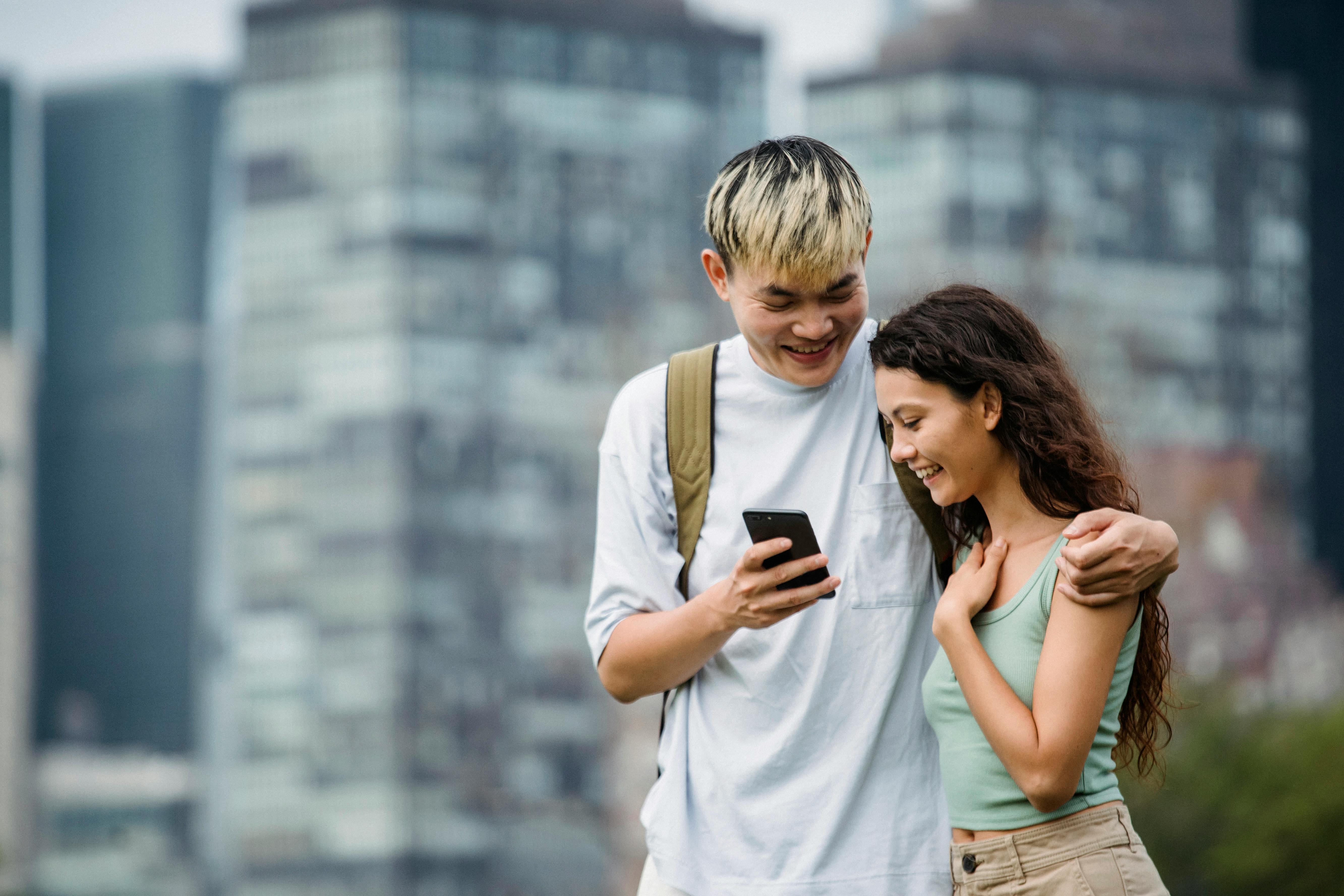 cheerful asian man showing smartphone to charming girlfriend in town