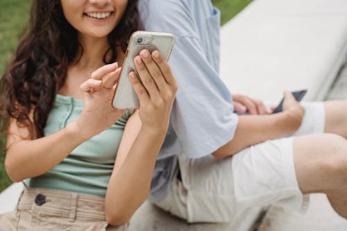 Free Crop young cheerful female browsing internet on cellphone while resting on bench near unrecognizable partner in town Stock Photo