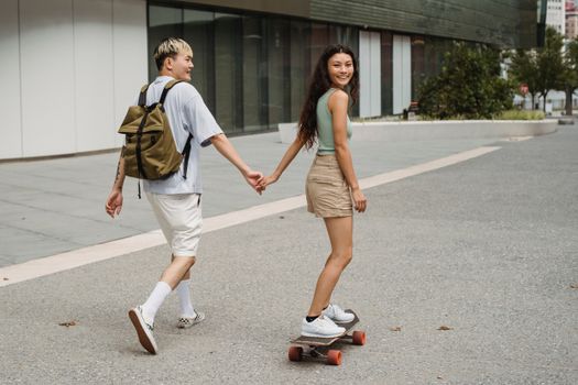 一名穿着休闲服、面带微笑的亚洲女子骑着滑板，牵着男友的手走在城市人行道上，看向镜头