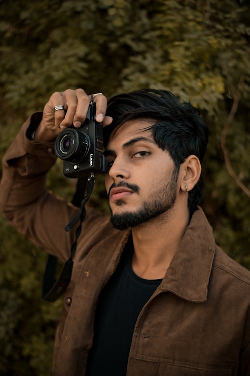Confident young ethnic man with black hair and beard in trendy clothes standing on street and shooting with photo camera and looking at camera at daytime