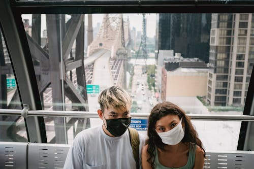 Young couple in casual outfit and protective face masks riding cableway cabin along urban New York City district near Queensboro Bridge during coronavirus outbreak