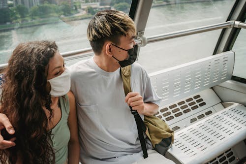 Positive couple wearing casual clothes and protective face masks embracing and riding ropeway cabin above contemporary city river