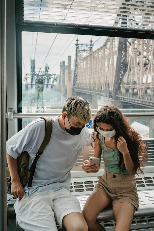 Focused young couple in  casual outfit and protective face masks riding cableway cabin together in modern city and surfing mobile phone