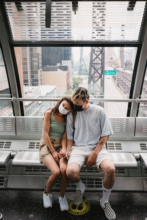 Full body young couple in casual clothes and protective masks riding ropeway observation cabin together while traveling in urban metropolis city during coronavirus epidemic