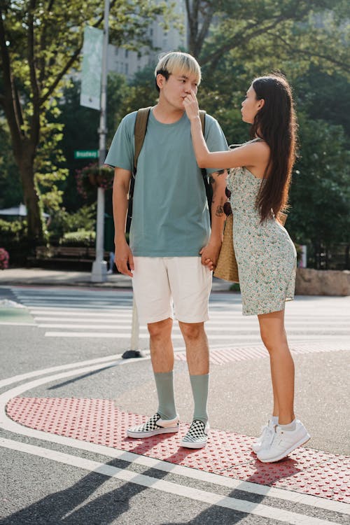 Full body of gentle loving girlfriend standing on tiptoes and touching face of ethnic boyfriend while standing on crosswalk
