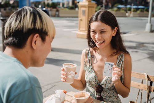 Cheerful ethnic female with cup of coffee showing photo made on instant photo camera while having lunch with boyfriend