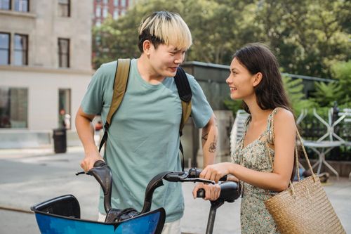 Free Cheerful multiethnic couple looking at each other and standing with bicycles on street with green trees Stock Photo