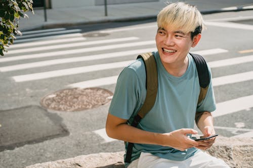 Cheerful Asian male with trendy hairstyle texting message on mobile phone and looking away on street with pedestrian cross