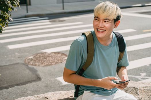一位发型时尚的开朗亚洲男性，在人行横道上用手机发短信，目光看向别处