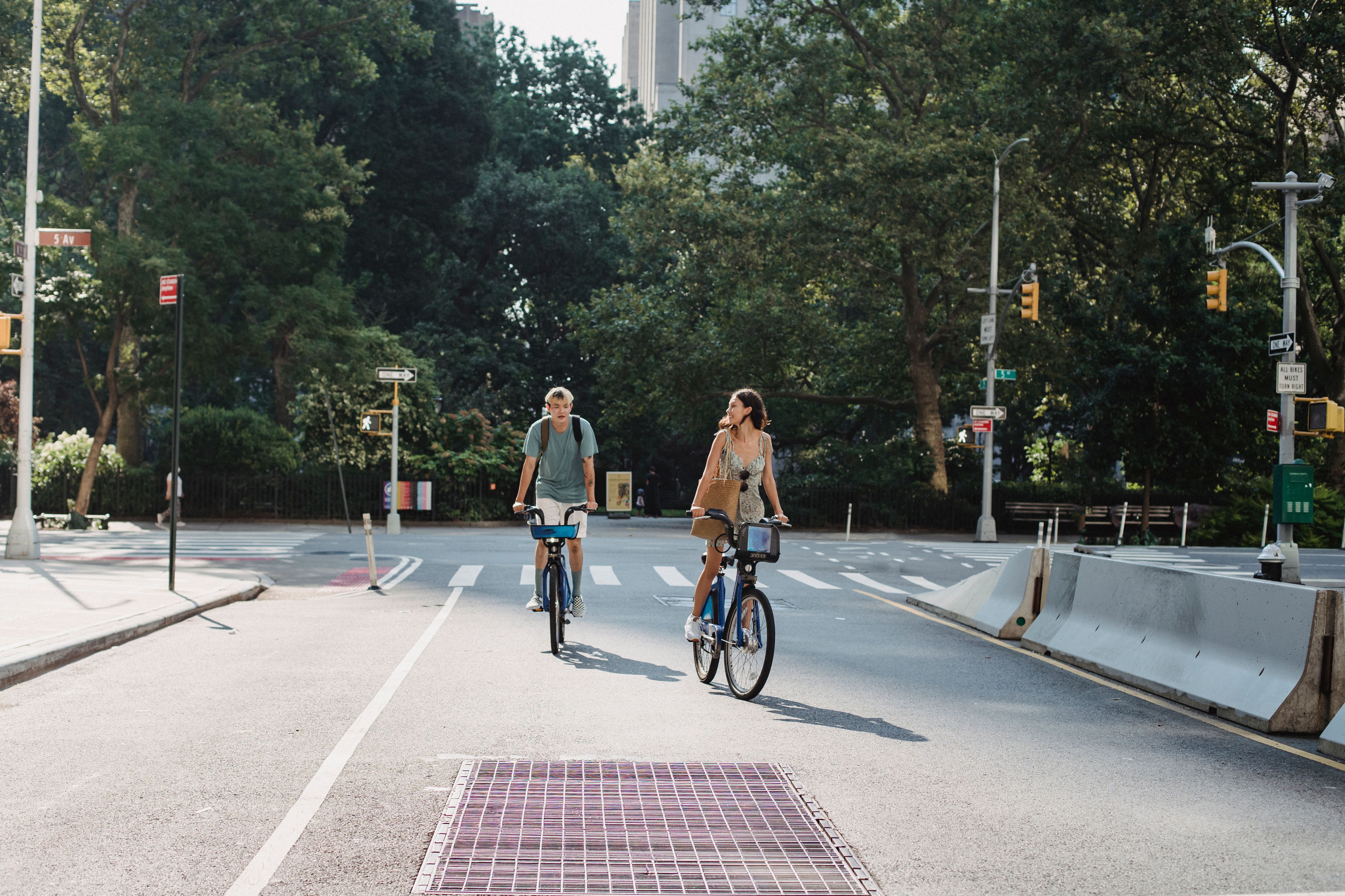 Couple riding bicycles on asphalt roadway in city against green trees in sunny day