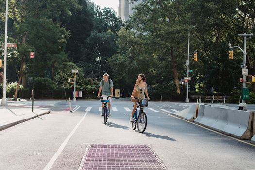 Couple riding bicycles on asphalt roadway in city against green trees in sunny day