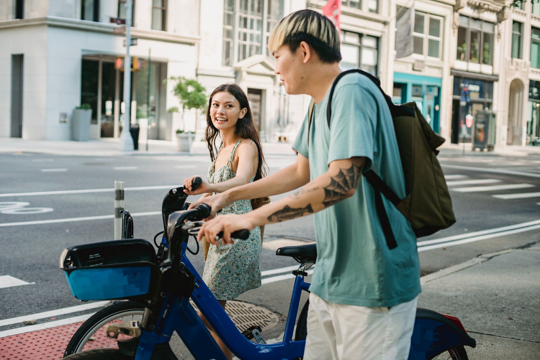 Smiling ethnic female in summer dress riding on bicycle with friend in city in daytime