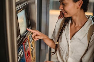 Crop smiling Asian female in white shirt using ticket vending machine with touch screen in underground