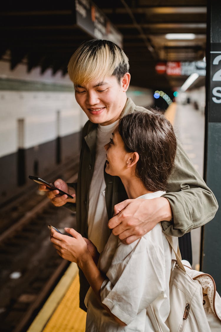 Happy Young Ethnic Couple Standing On Platform Of Underground Station With Smartphones