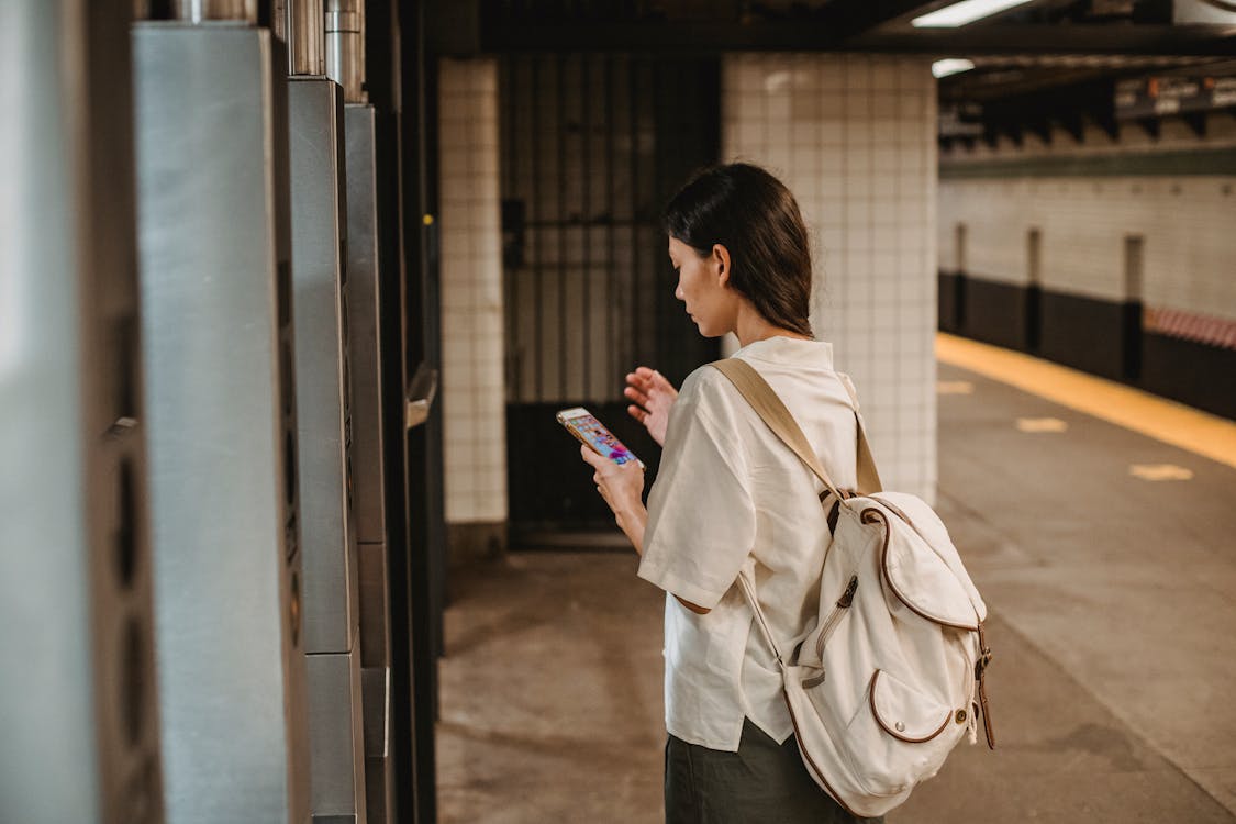 Side view of young concentrated ethnic female in casual clothes and backpack browsing mobile phone while standing on platform of subway station