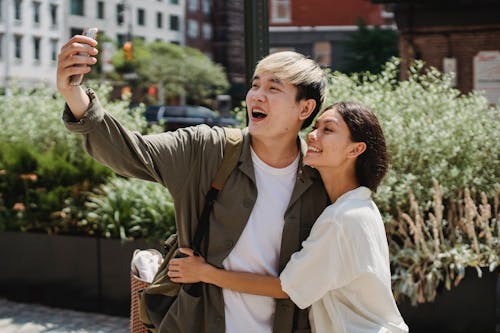 Free Cheerful young Asian couple in stylish clothes cuddling and smiling while taking selfie on mobile phone during date in city Stock Photo