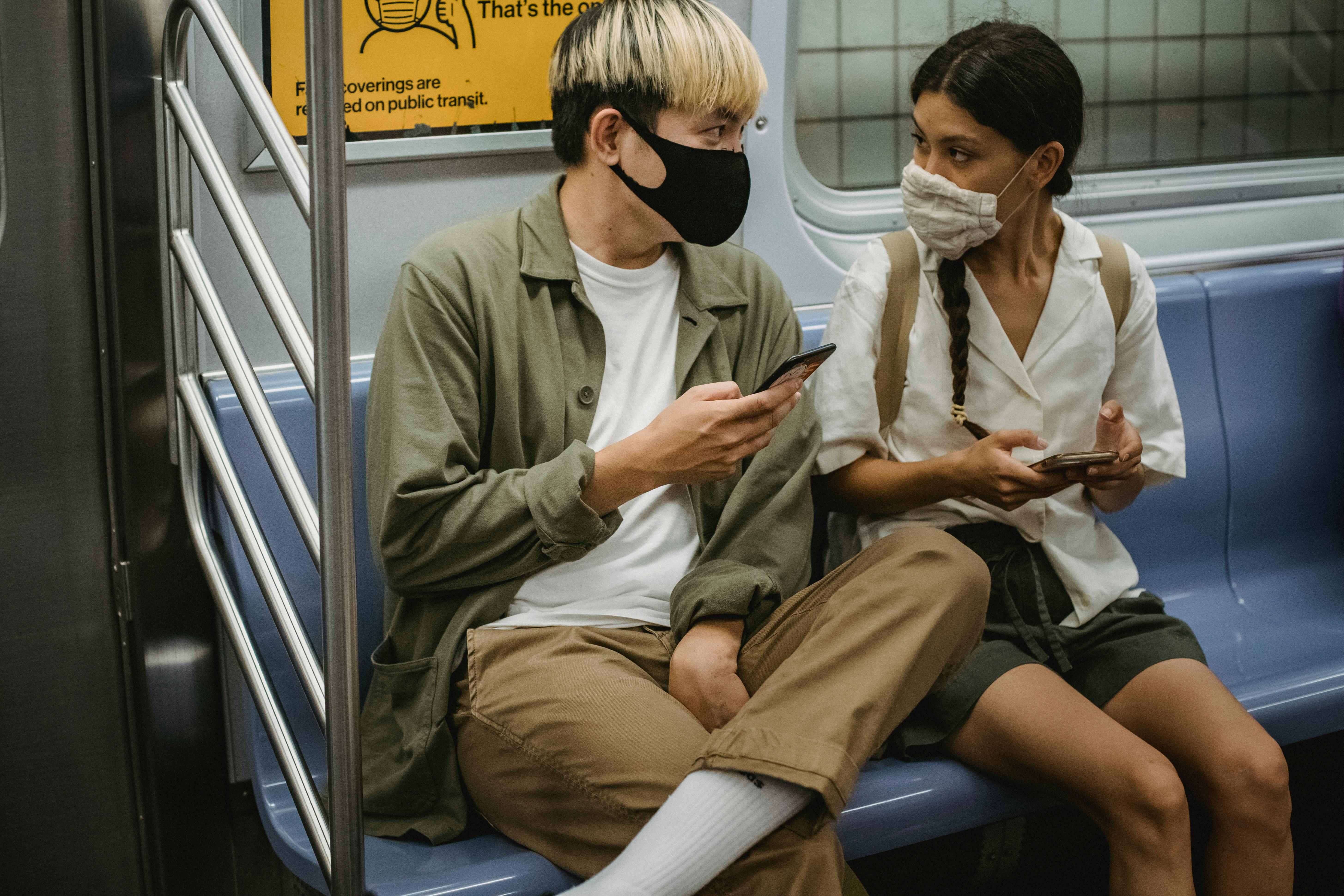 young diverse couple in medical masks riding train and browsing smartphones