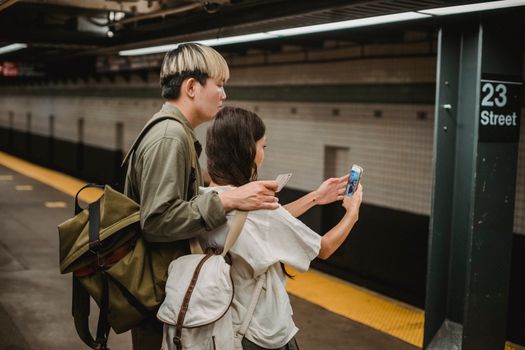 Side view of ethnic young female in casual clothes and backpack taking selfie with Asian boyfriend on smartphone while standing on platform in subway station