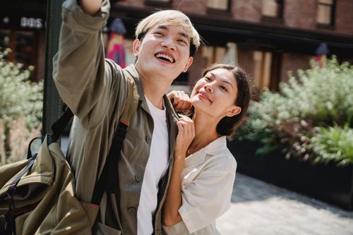 Free Cheerful young Asian couple in trendy outfits cuddling and smiling while taking selfie on street during romantic holidays on sunny day Stock Photo