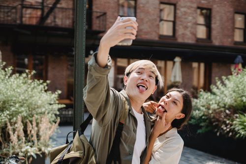 Free Playful young Asian couple showing tongue and taking selfie on smartphone while having fun on street on sunny day Stock Photo