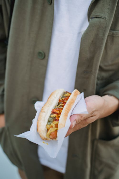 From above anonymous male in casual clothes holding yummy hot dog on napkin while standing on street