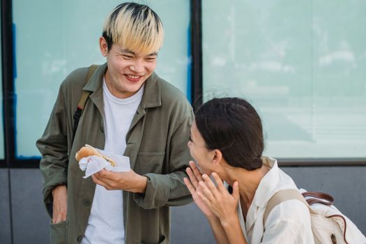 Delighted young Asian couple eating yummy hot dog on sunny street while looking at each other and laughing