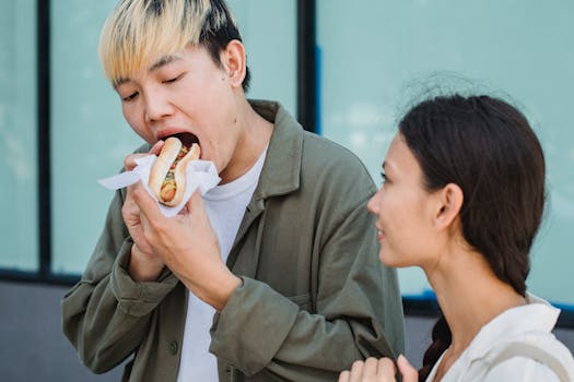 Content Asian male in casual outfit eating delicious hot dog while standing on sunny street near attractive girlfriend