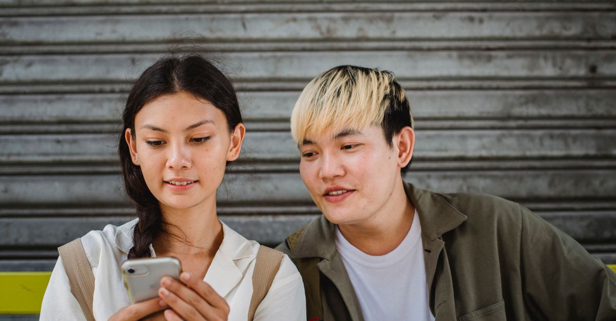 Content young Asian couple in casual outfits surfing contemporary mobile phones and sitting on street bench on clear weather