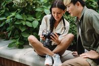 Crop glad Asian couple wearing summer clothes sitting on park parapet and looking through photos on contemporary photo camera