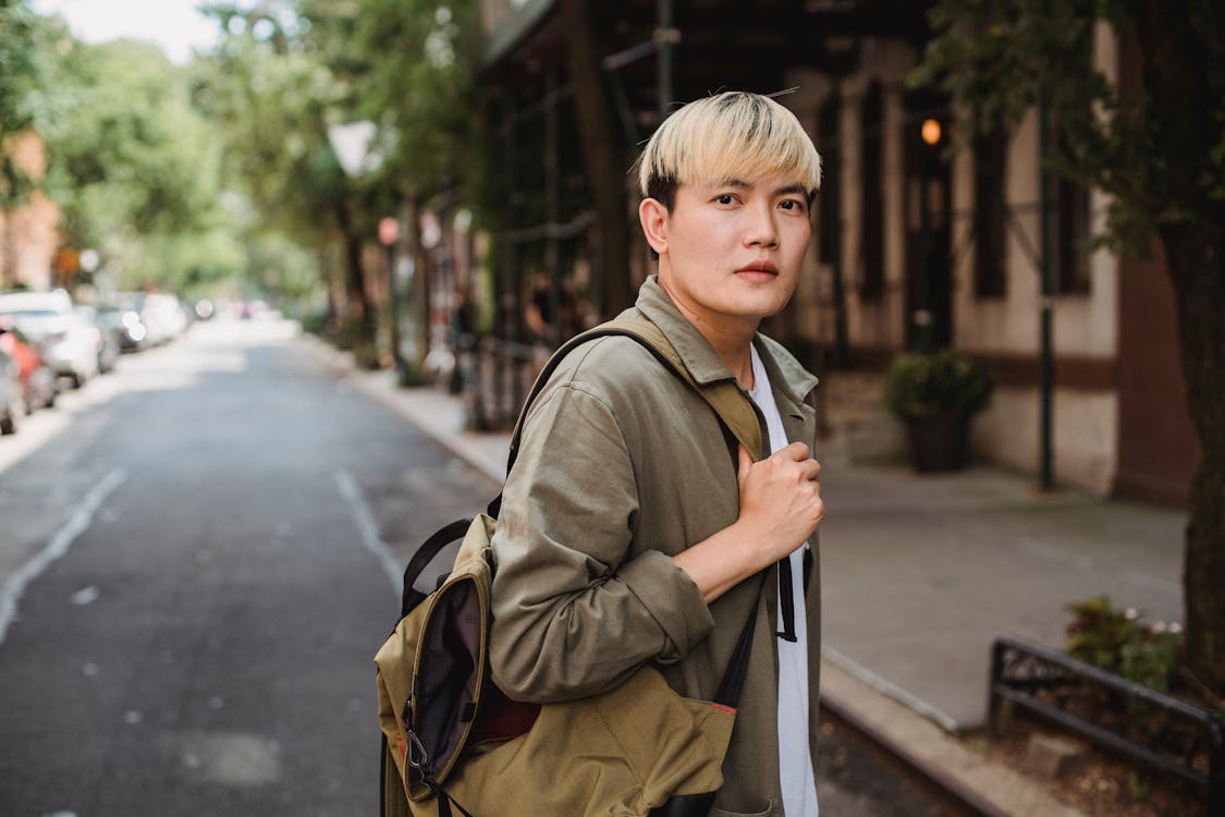 Side view emotionless young Asian male in casual clothes standing with backpack on shoulder on sunny town street and looking at camera
