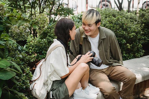 Young content multiracial couple with photo camera interacting on bench near shrubs while looking at each other