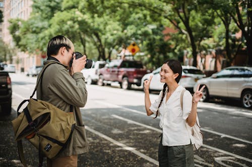 Free Anonymous male with rucksack taking photo of cheerful ethnic beloved showing peace gesture on urban roadway Stock Photo