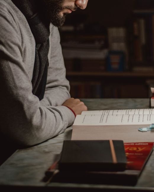Man reading at a desk in a dimly lit room with textbooks and notes, focused on learning. Man reading at a desk in a dimly lit room with textbooks and notes, focused on learning.