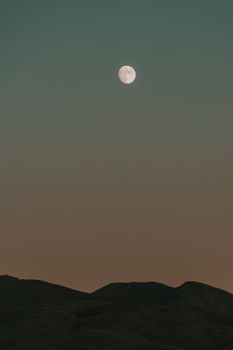 A tranquil night scene featuring a full moon illuminating mountains under a clear night sky.