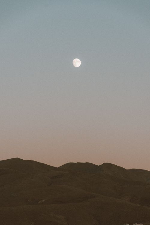 Free A serene image capturing a full moon rising over silhouetted desert hills at dusk. Stock Photo
