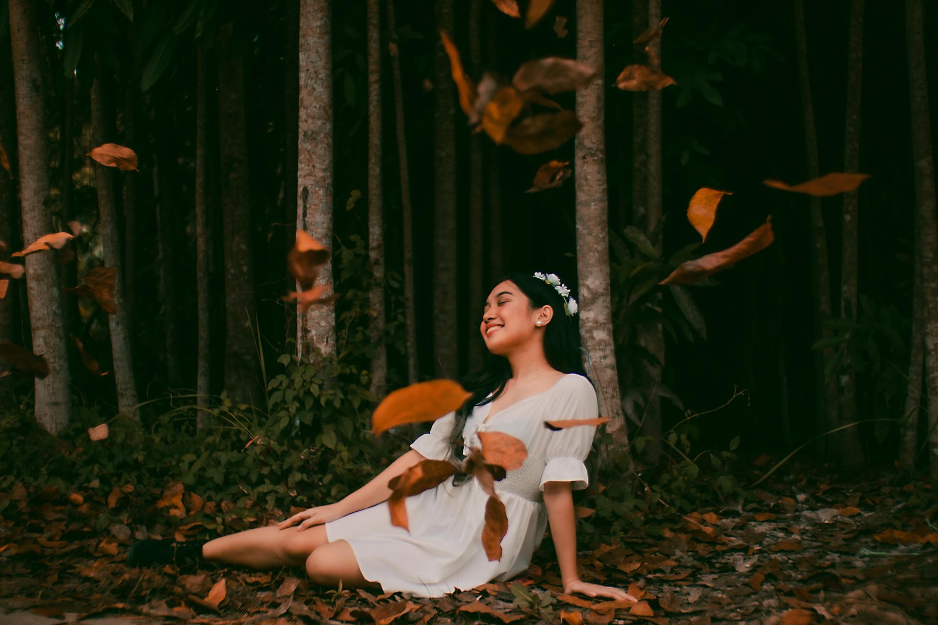 Woman in White Dress Sitting on Ground While Leaves Fall
