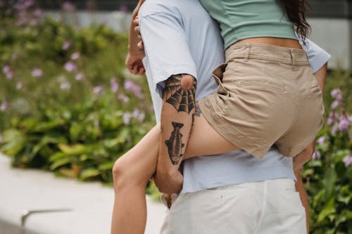 Back view of crop faceless young tattooed guy giving piggyback ride to slim girlfriend while spending time together in park on sunny day