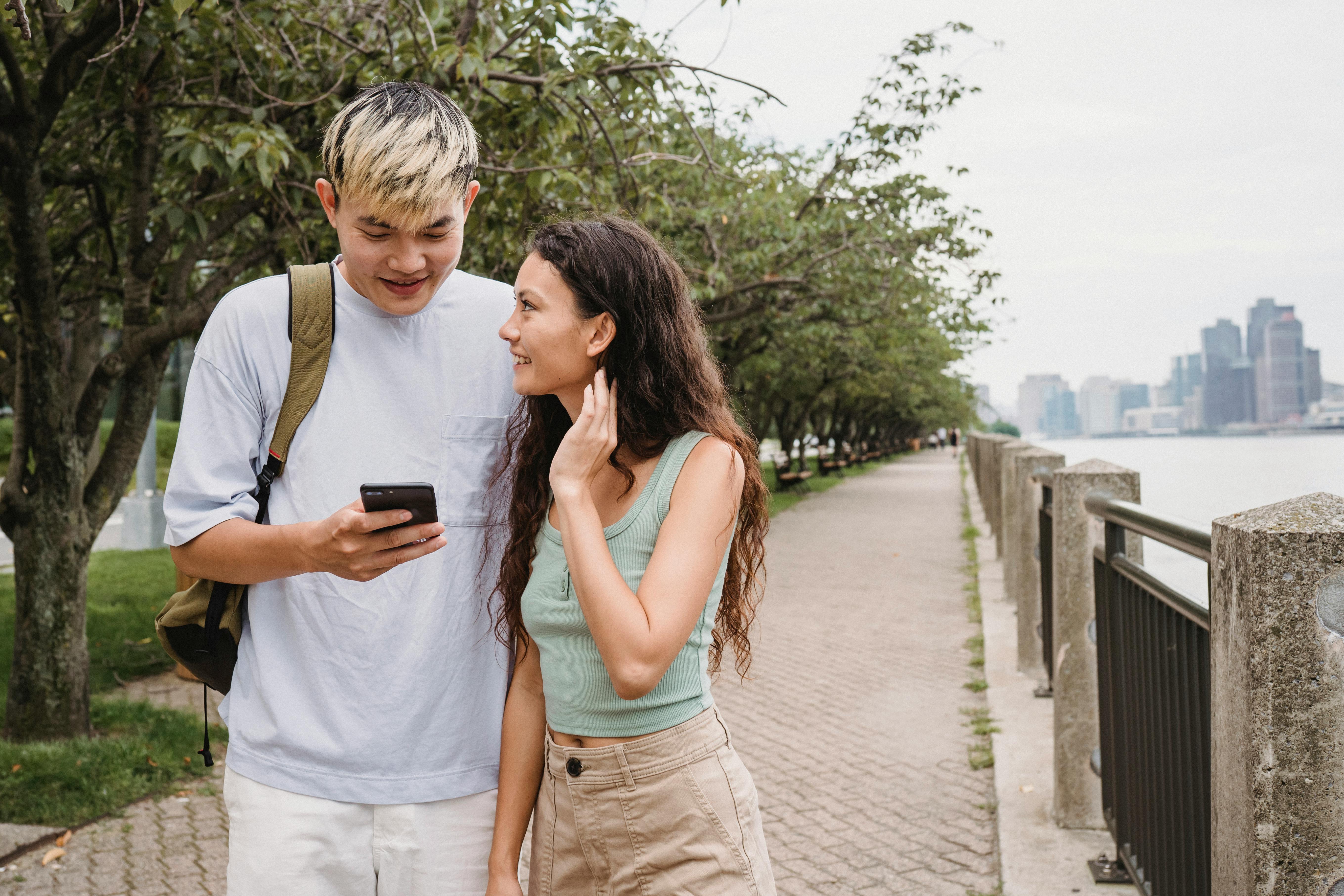 positive young ethnic couple using smartphone while strolling in city park