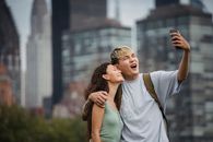 Cheerful young ethnic couple hugging and making face while standing in park near modern skyscrapers and taking selfie on smartphone