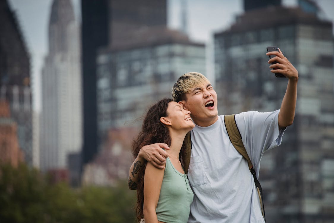 Cheerful young ethnic couple hugging and making face while standing in park near modern skyscrapers and taking selfie on smartphone