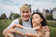 Cheerful Asian boyfriend hugging ethnic girlfriend making grimace while taking self portrait on cellphone in green city park