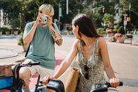 Anonymous male with instant camera taking picture of unrecognizable girlfriend on bicycle while standing on pavement on street in city