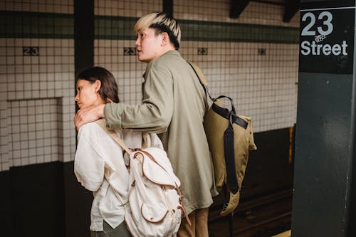 Free Back view of Asian couple with backpacks hugging and standing on platform of subway station and leaning forward while waiting for train Stock Photo
