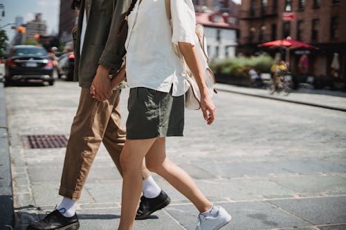 Free Side view of unrecognizable couple holding hands and crossing road on sunny street in city with blurred cars and buildings Stock Photo