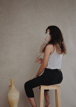 一位身着时尚服装、面目全非的女士，脸上沾满蓬松的植物，正坐在陶罐附近的木凳上