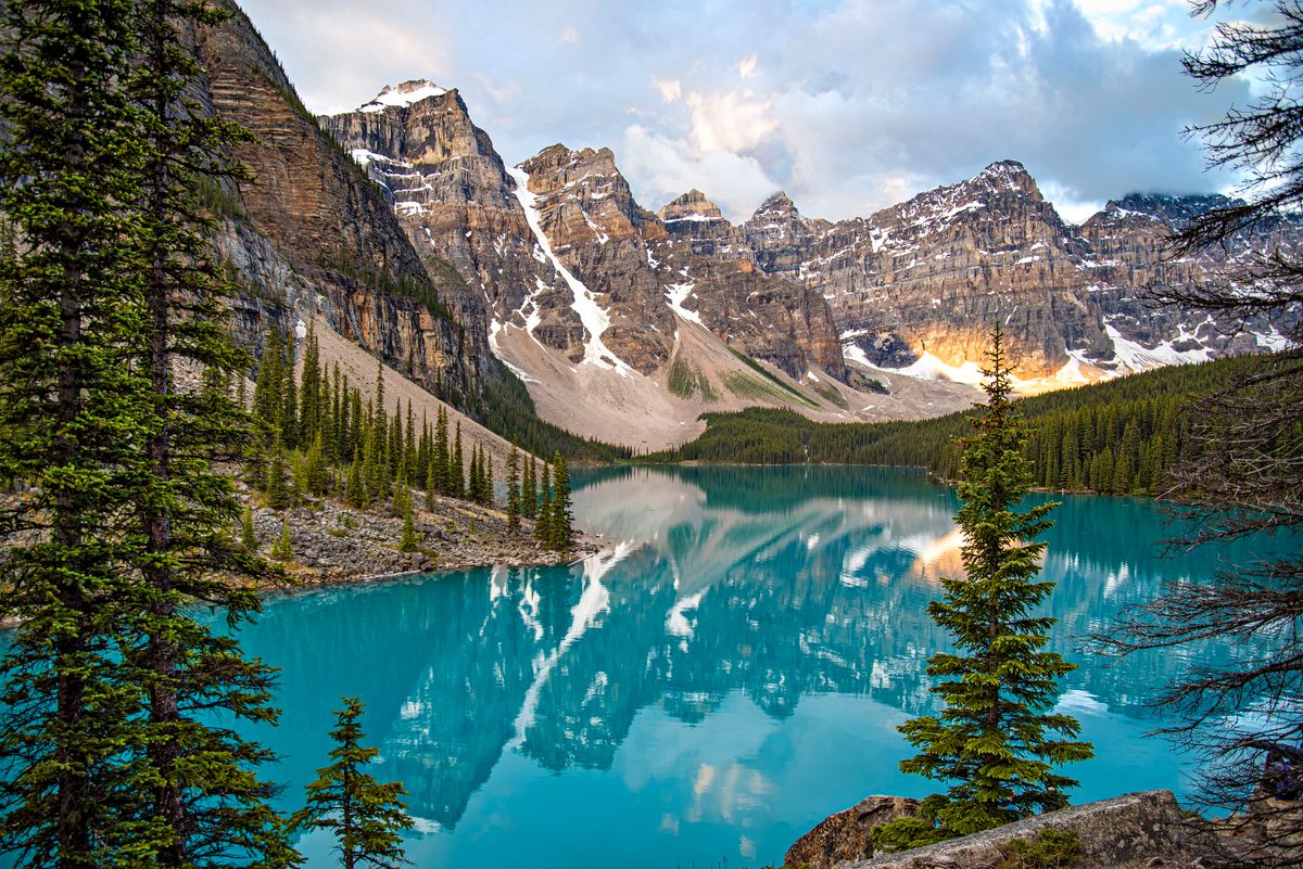 Breathtaking view of Moraine Lake with reflection of snow-covered mountains, pine trees, and vibrant blue water.