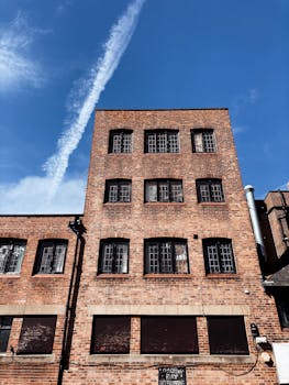 From below of long white contrail from airplane in vivid blue sky over tall multi storey building constructed of bricks