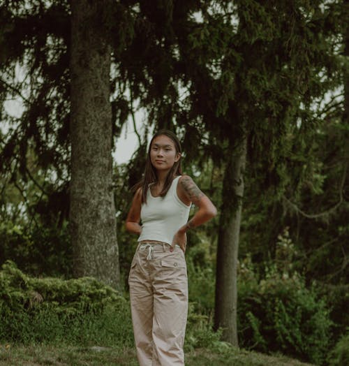 Smiling Asian female with hands on waist standing in green woodland and enjoying nature