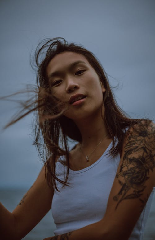 Free Low angle of Asian female with tattoo and flying hair resting in nature near sea in  cloudy day Stock Photo